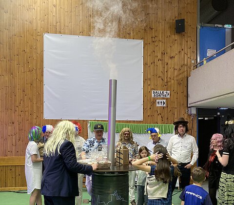 Menschen in Kostümen und Alltagskleidung stehen um einen Metallofen mit Rauch in einer Halle mit Holzwänden.
