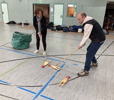 Zwei Personen spielen in einer Halle mit Besen und Gummihühnern auf dem Boden.