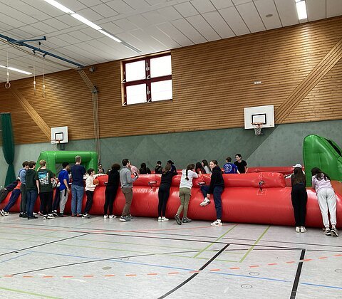 Gruppe von Jugendlichen spielt auf aufblasbarem Menschenkicker in einer Sporthalle mit Holzwänden und Basketballkörben.