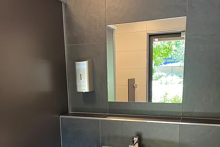 Waschbecken mit Spiegel, Seifenspender und Mülleimer in einem modernen, dunklen Badezimmer mit Fensterblick.