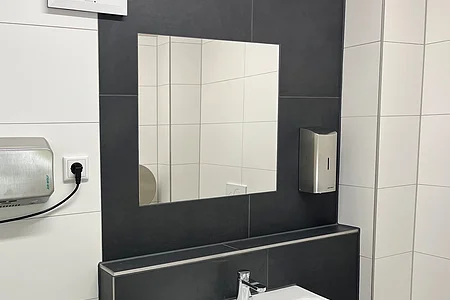 Waschbecken mit Spiegel, Seifenspender und Handtrockner in einem modernen, schwarz-weiß gefliesten Badezimmer.