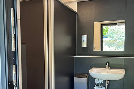 Barrierefreies WC mit grauen Trennwänden, Waschbecken, Spiegel, Seifenspender und Mülleimer.