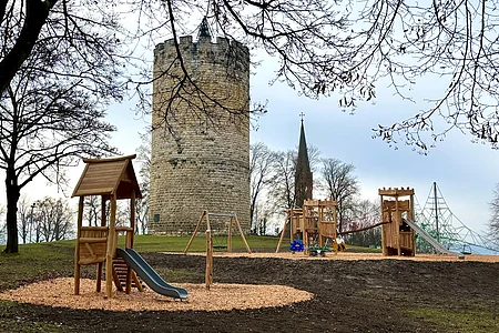 Spielplatz mit Rutsche und Schaukeln vor einem runden Steinturm und Kirchturm im Hintergrund, kahle Bäume.