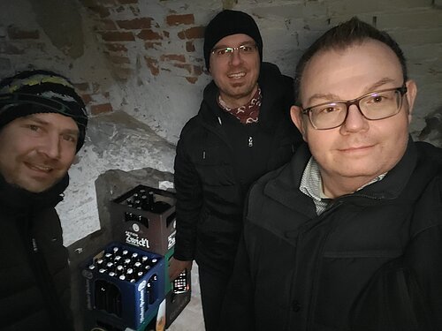 Drei Männer in Winterkleidung in einem Kellerraum mit Bierkästen im Hintergrund.