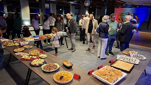 Mehrere Personen stehen in einem Raum mit Tischen, auf denen verschiedene Speisen als Buffet angerichtet sind.