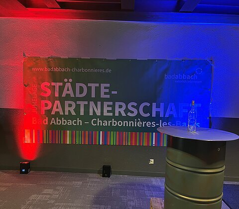 Banner mit Schrift „Städtepartnerschaft Bad Abbach – Charbonnières-les-Bains“ an Wand mit roter und blauer Beleuchtung.