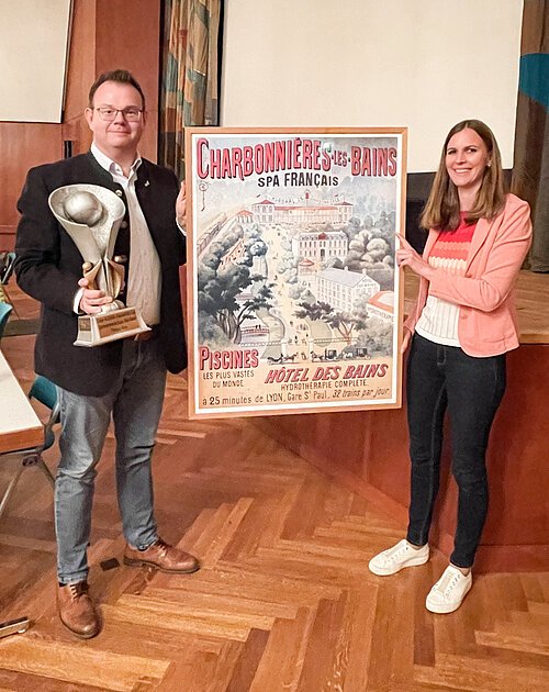 Mann mit Pokal und Frau halten ein großes Plakat mit französischem Text in einem Raum mit Holzfußboden.