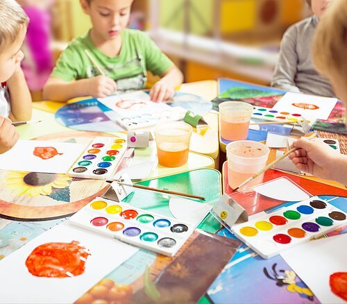 Kinder sitzen an einem Tisch und malen mit Wasserfarben auf Papier in einem Klassenraum.