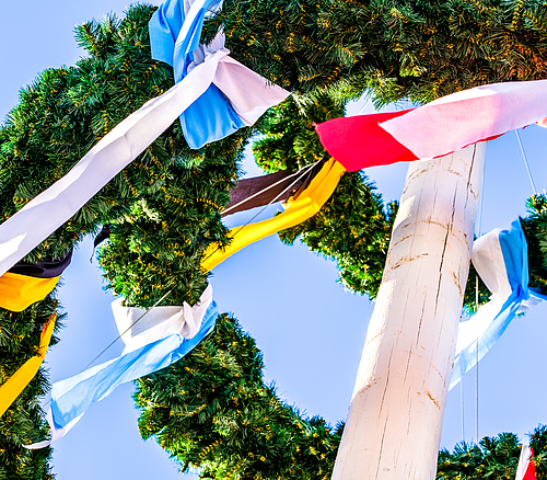 Maikranz mit bunten Bändern an einem weißen Maibaum vor blauem Himmel, Blick nach oben.