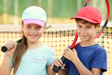 Zwei Kinder mit Tennisschlägern stehen vor einem Tennisnetz auf einem Tennisplatz.