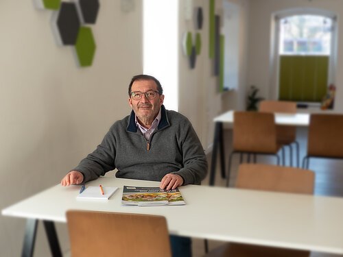 Älterer Mann mit Brille sitzt an weißem Tisch mit Heft und Stiften in modernem Raum mit Stühlen und Fenster.
