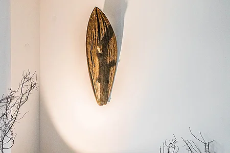 Holzskulptur an weißer Wand mit Schatten, links und rechts kahle Zweige sichtbar