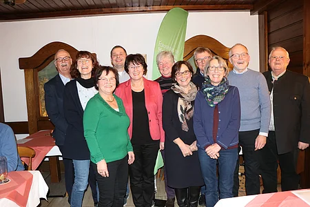Elf Personen stehen in einem Raum, lächeln in die Kamera, im Hintergrund ein grünes Banner.