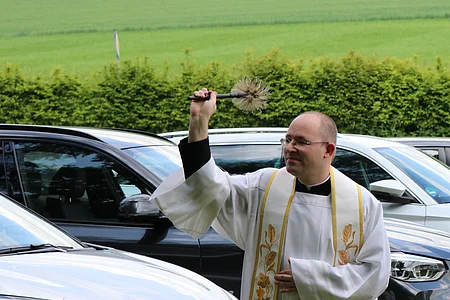 Priester in weißem Gewand segnet parkende Autos mit Weihwasser im Freien auf einer Wiese.