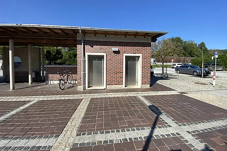 Zwei nebeneinanderliegende öffentliche Toiletten mit Metalltüren an einem Backsteingebäude bei sonnigem Wetter.