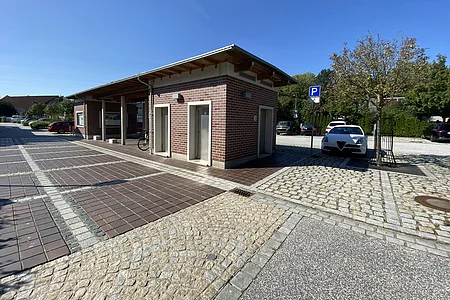 Gebäude mit drei Türen auf gepflastertem Platz bei klarem Himmel und geparktem Auto neben Baum.