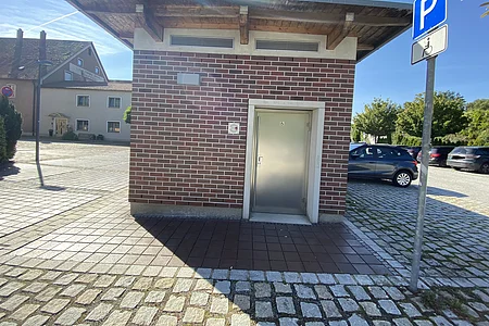 Kleines Gebäude mit Metalltür und Rollstuhlsymbol, daneben Parkplatz mit Behindertenparkplatzschild.