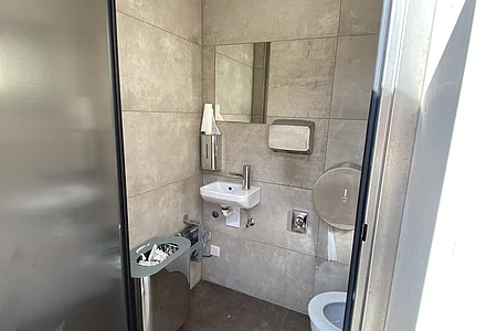 Kleines WC mit grauen Fliesen, kleinem Waschbecken, Spiegel, Papierhandtuchspender, Mülleimer und Toilettenpapierhalter.