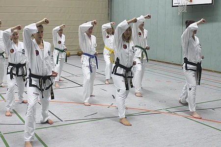Gruppe von Menschen in weißen Kampfsportanzügen mit farbigen Gürteln beim Training in einer Halle.