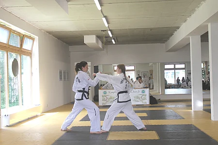Zwei Frauen in weißen Taekwondo-Anzügen mit schwarzen Gürteln trainieren in einem Dojo.