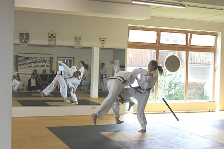 Zwei Personen in weißen Kampfanzügen führen einen Wurf in einem Trainingsraum mit Spiegel und großen Fenstern aus.