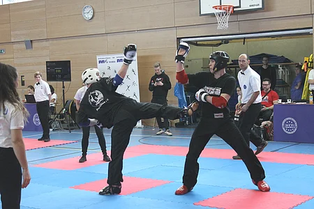 Zwei Kampfsportler mit Schutzhelm und Handschuhen kämpfen auf einer Matte in einer Halle, Schiedsrichter und Zuschauer im Hintergrund.