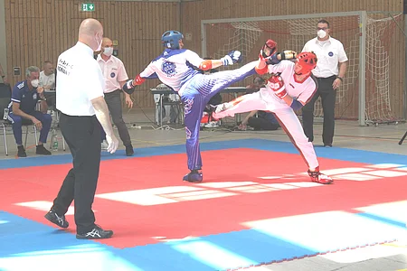 Zwei Kampfsportler mit Schutzhelm und Handschuhen führen einen Kick auf einer roten Matte in einer Halle aus.