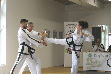 Drei Personen in Taekwondo-Anzügen, eine Frau führt einen Tritt zum Brett, das zwei Männer halten.
