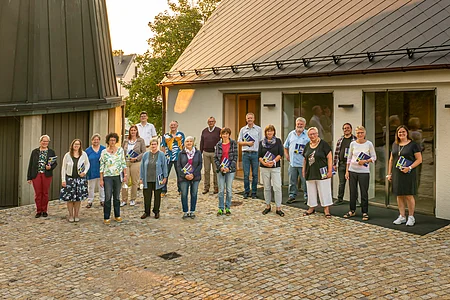 Gruppenfoto von 18 Personen vor einem Gebäude mit Schieferdach, alle halten Broschüren in der Hand.