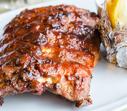 Nahaufnahme von gegrillten Rippchen mit Barbecue-Sauce auf weißem Teller, daneben Folienkartoffel mit Butter.
