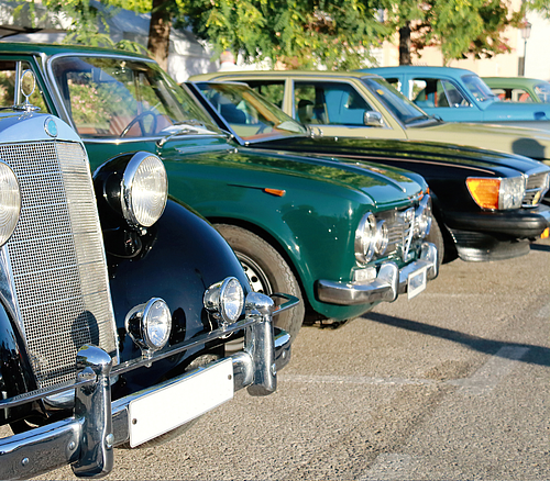 Vorderansicht von vier geparkten Oldtimer-Autos in verschiedenen Farben auf einem sonnigen Parkplatz.