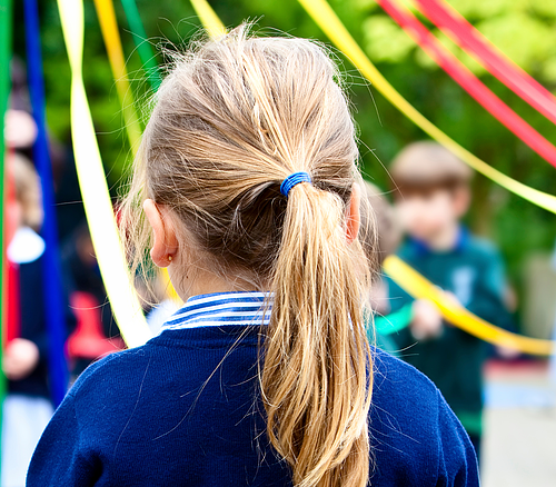 Kind mit Pferdeschwanz von hinten, umgeben von bunten Bändern im Freien, weitere Kinder unscharf im Hintergrund.