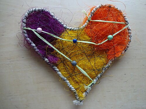 Herzförmiges Kunstobjekt aus buntem Draht und Fasern in Lila, Gelb und Orange auf hellem Untergrund.
