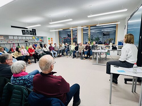 Gruppe von Menschen sitzt im Stuhlkreis in einem hellen Raum, eine Person spricht von einem Tisch aus.