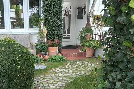 Eingang eines Hauses mit weißer Tür, roten Dachüberstand, Pflanzen und gepflastertem Weg im Vorgarten.