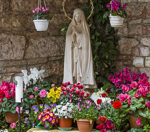 Statue der betenden Maria mit Rosenkranz, umgeben von bunten Blumen und zwei hängenden Blumentöpfen vor Steinwand.