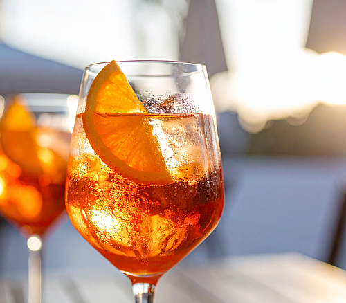 Glas mit orangenfarbenem Getränk, Eiswürfeln und Orangenscheibe auf Holztisch bei Sonnenlicht.