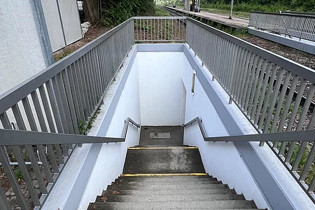 Treppe mit Geländer führt von Bahnsteig zu Unterführung an einem Bahnhof mit Gleisen und Bäumen im Hintergrund.