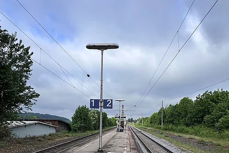 Bahnsteig mit zwei Gleisen, Lampen und Schildern mit den Nummern 1 und 2, bewölkter Himmel, Bäume links und rechts.