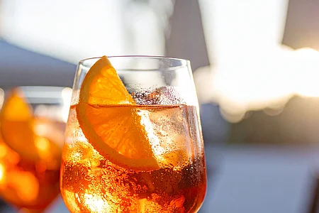 Glas mit orangenfarbenem Getränk, Eiswürfeln und Orangenscheibe auf Holztisch bei Sonnenlicht.