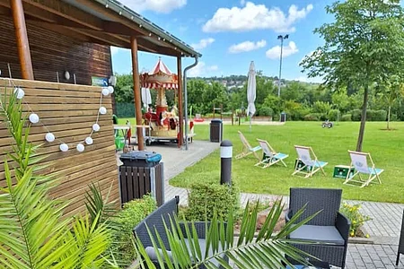 Außenbereich mit grauen Sitzmöbeln, Pflanze im Vordergrund und Liegestühlen auf einer Wiese im Hintergrund.