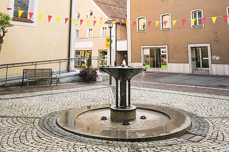 Kleiner Platz mit Brunnen, umgeben von historischen Gebäuden, mit bunten Wimpeln geschmückt, unter bewölktem Himmel.