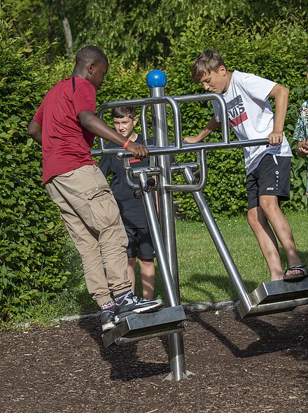 Fünf Jungen spielen an einem Outdoor-Fitnessgerät auf einem Spielplatz mit grünem Hintergrund.