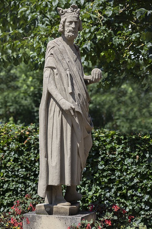 Steinerne Statue eines bärtigen Königs mit Krone und Kugel in der rechten Hand umgeben von grünen Pflanzen.