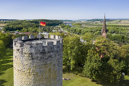 Runder Steinturm mit deutscher Flagge neben Kirchturm in grüner Landschaft mit Fluss und Feldern im Hintergrund.