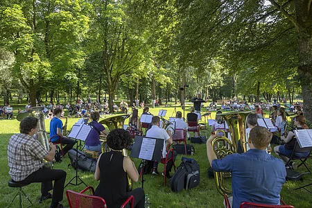 Orchester spielt im Park vor Publikum auf Wiesenfläche unter Bäumen bei Tageslicht
