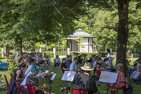 Orchester mit Blasinstrumenten spielt im Park unter Bäumen, Zuschauer sitzen im Hintergrund auf Bänken.