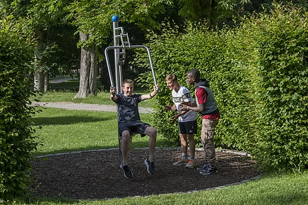 Drei Jungen im Park, einer sitzt an einem Outdoor-Fitnessgerät, zwei stehen daneben und schauen zu.