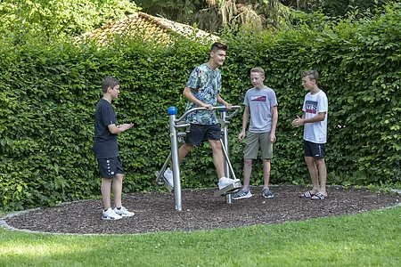 Vier Jungen stehen auf einem Spielplatz, einer benutzt ein Outdoor-Fitnessgerät, umgeben von Hecke und Rasen.
