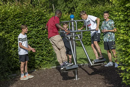 Fünf Jungen spielen an einem Outdoor-Fitnessgerät auf einem Spielplatz mit grünem Hintergrund.